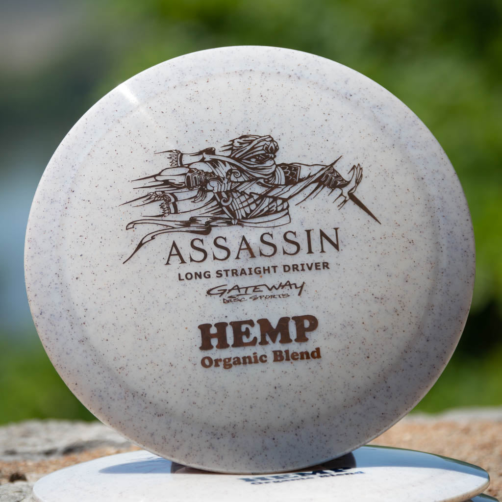 The Assassin - Diamond Hemp is a white speckled distance driver by Gateway Disc Sports, featuring assassin artwork and "ASSASSIN Long Straight Driver Diamond Hemp Blend" text, known for its neutral flight path.