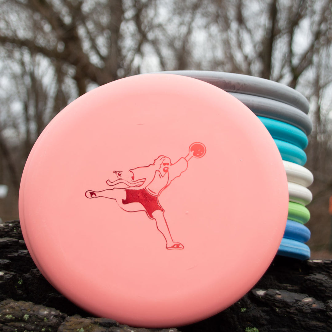 Outdoors, a stack of colorful disc golf discs is shown, featuring the Wizard 10 Pack - Airdude Variety Pack with a pink putter in the foreground decorated with artwork of a person throwing a disc.