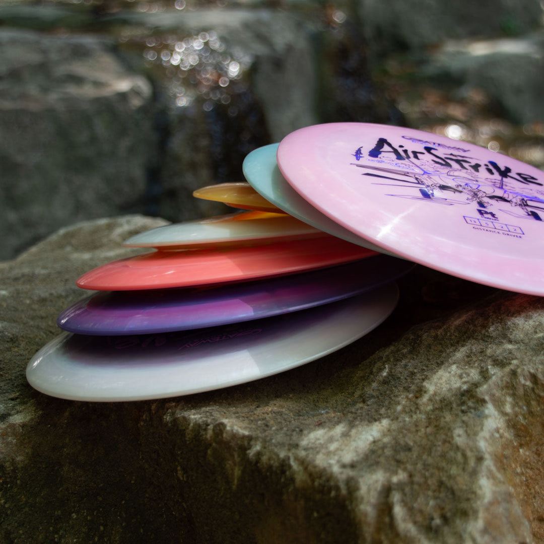 A stack of Airstrike - Platinum long range disc golf drivers sits on a large rock outdoors, with water and rocks in the background.