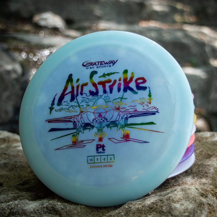 A light blue Airstrike - Platinum long range driver disc rests on a rock, featuring a colorful airplane graphic, great glide, easy control, and flight numbers 13 | 5 | -2 | 2.