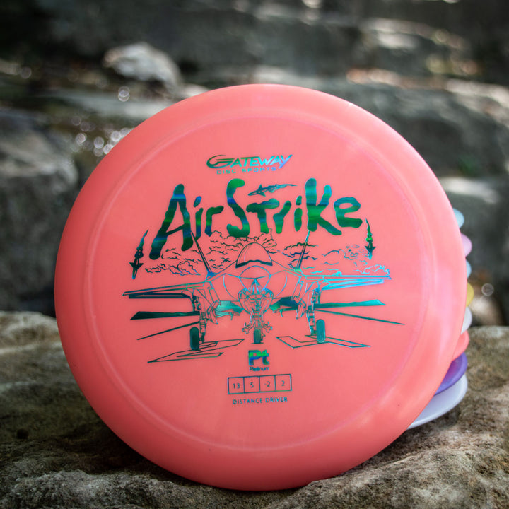 The Airstrike - Platinum is a pink disc golf driver with a fighter jet illustration, renowned for its excellent glide, shown resting on an outdoor rock.