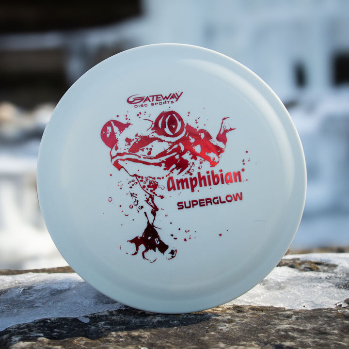 A white disc golf disc featuring a red frog illustration and "Amphibian - SureGrip™ Superglow" text, this smooth control driver from Gateway Disc Sports rests on a rock, ready for your next round.