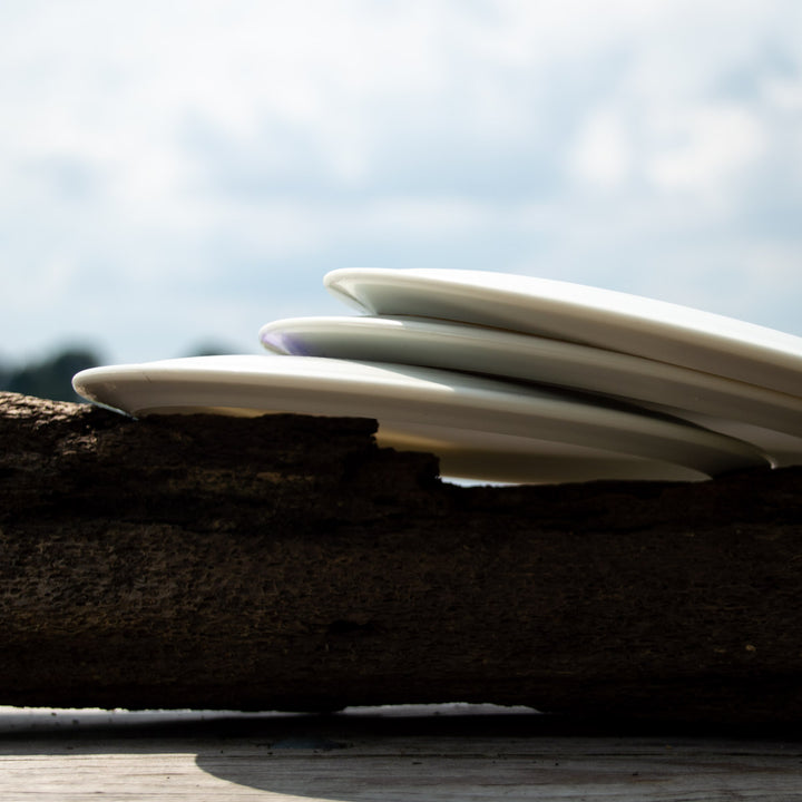 Four Amphibian - SureGrip™ Superglow discs, stacked on a rustic wooden surface, resemble classic disc golf gear against a softly blurred outdoor backdrop.
