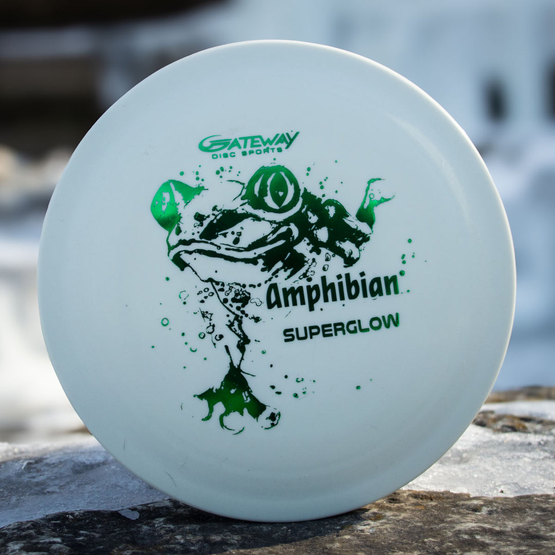 A white disc golf disc with a green frog and "Gateway Disc Sports Amphibian - SureGrip™ Superglow" text rests on a rock, ideal for players wanting a smooth control driver.