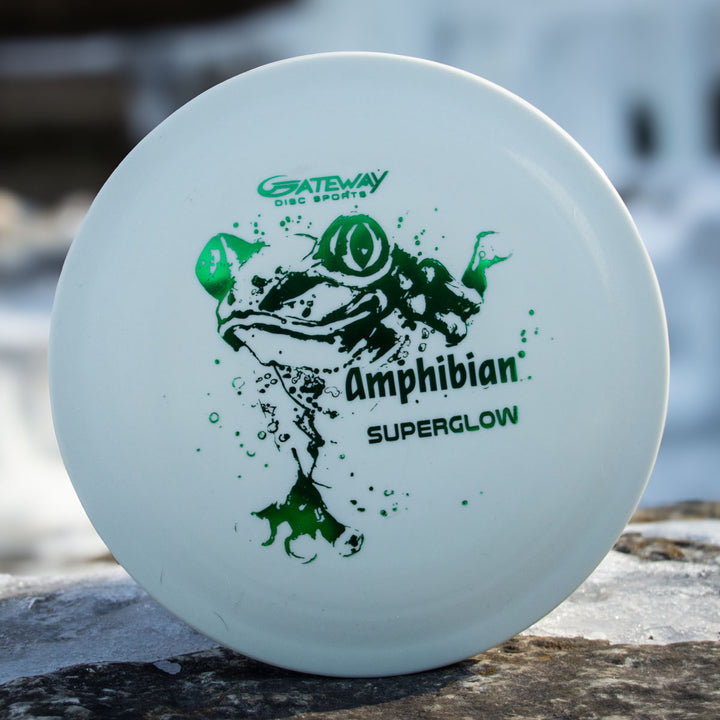 A white disc golf disc with a green frog and "Gateway Disc Sports Amphibian - SureGrip™ Superglow" text rests on a rock, ideal for players wanting a smooth control driver.