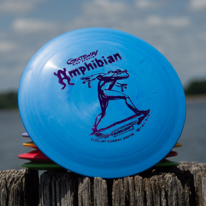 A blue Amphibian - SureGrip™ disc, ideal for controlled fairway drives and featuring a humanoid frog graphic, sits on a weathered wooden post near the water.