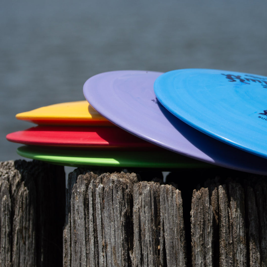 Four colorful frisbees, including the Amphibian - SureGrip™ known for smooth control driver performance, are stacked on a weathered wooden post beside a body of water.
