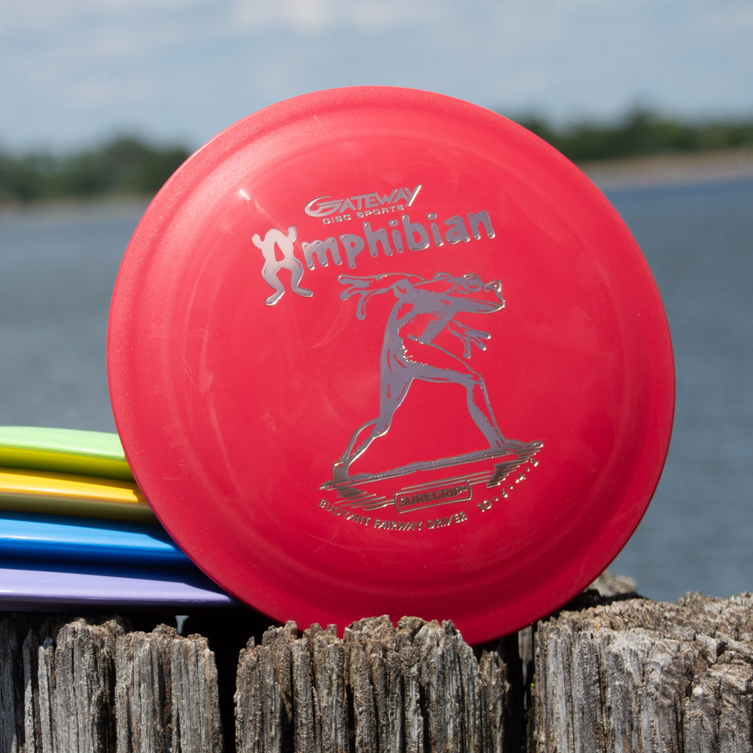 A red Amphibian - SureGrip™ disc featuring a frog graphic rests on a wooden post by water, with other colorful discs stacked nearby. Known for smooth control driver performance—ideal for fairway drives.