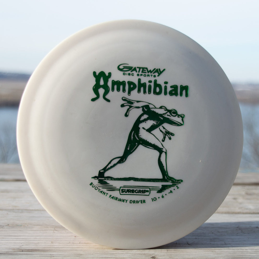 A white Amphibian - SureGrip™ disc, labeled "10•6•-1•2," shows a frog-like figure throwing a disc—ideal for smooth, controlled fairway drives.