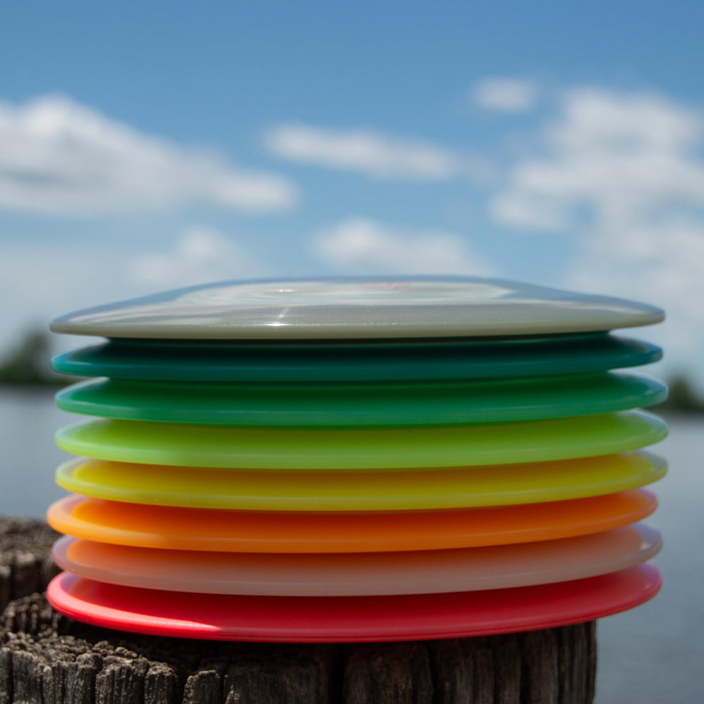 A stack of colorful disc golf discs, featuring the Apex - Diamond high-speed driver known for its stable flight path, sits outdoors on a wooden surface with a lake and blue sky in the background.