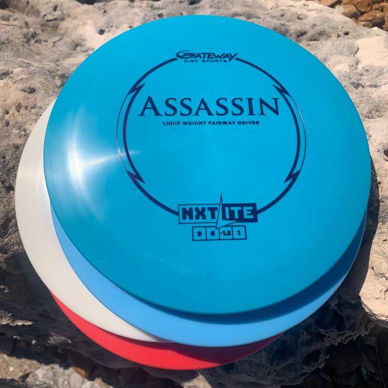 Three disc golf discs, with the top blue one labeled "Assassin - NXT LITE," a controllable distance driver known for excellent glide, stacked on a large rock outdoors.