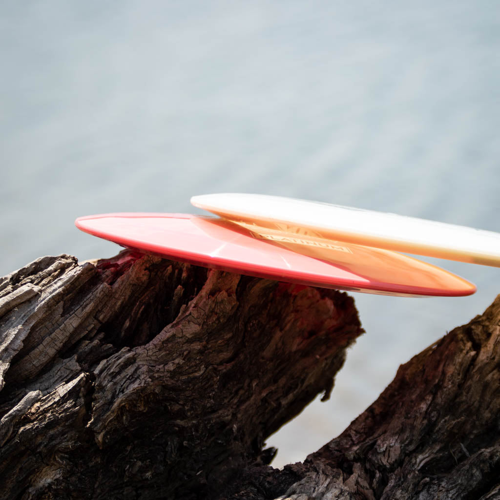 Two vibrant Assassin - Platinum frisbees, one red and one white, lie on weathered driftwood by the water—both offer excellent glide for a controllable distance driver.