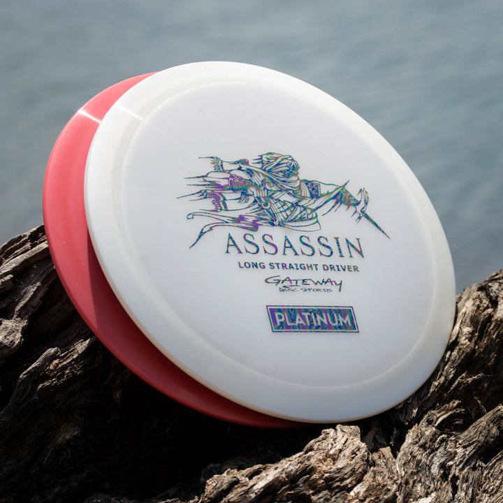 Two disc golf discs, one white with "Assassin Long Straight Driver" text and art, and one red, rest on wood by the water—ideal for players wanting a controllable distance driver with excellent glide like the Assassin - Platinum.