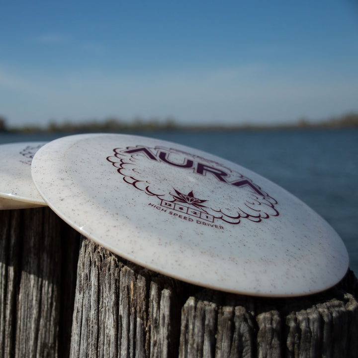 A white Aura - Diamond Hemp disc golf driver—renowned for easy turnover and long flights—rests on a weathered wooden post beside water beneath a clear blue sky.