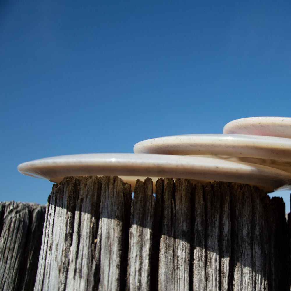 Three Aura - Diamond Hemp discs, reminiscent of white ceramic plates, are stacked on a weathered wooden fence post against a clear blue sky, embodying the simple elegance of easy turnover design.