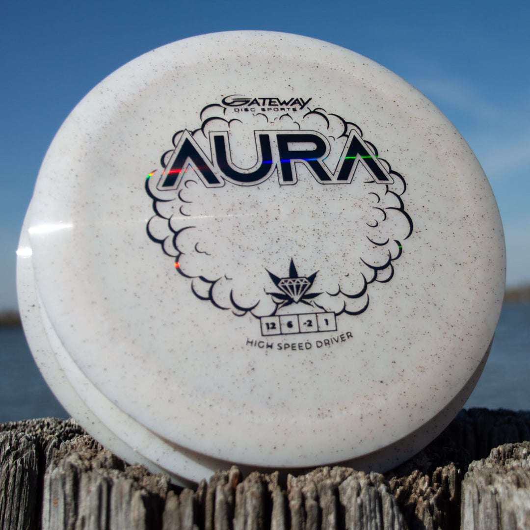 A white Aura - Diamond Hemp high speed driver sits on a wooden post outdoors with water and a blue sky in the background.