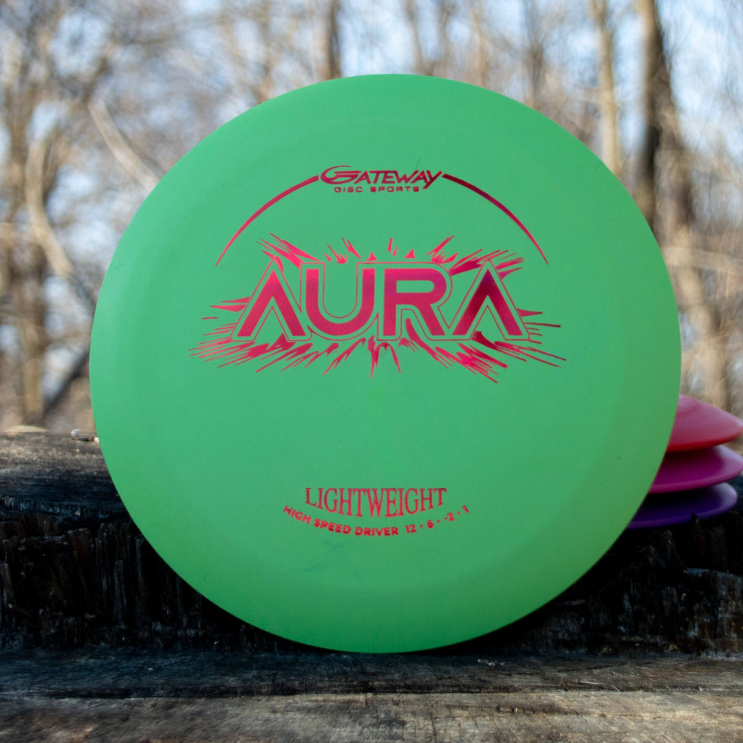 A green Aura - Suregrip Lightweight high-speed driver disc is shown outdoors on wood with trees behind it, designed for easy turnover and full flights.