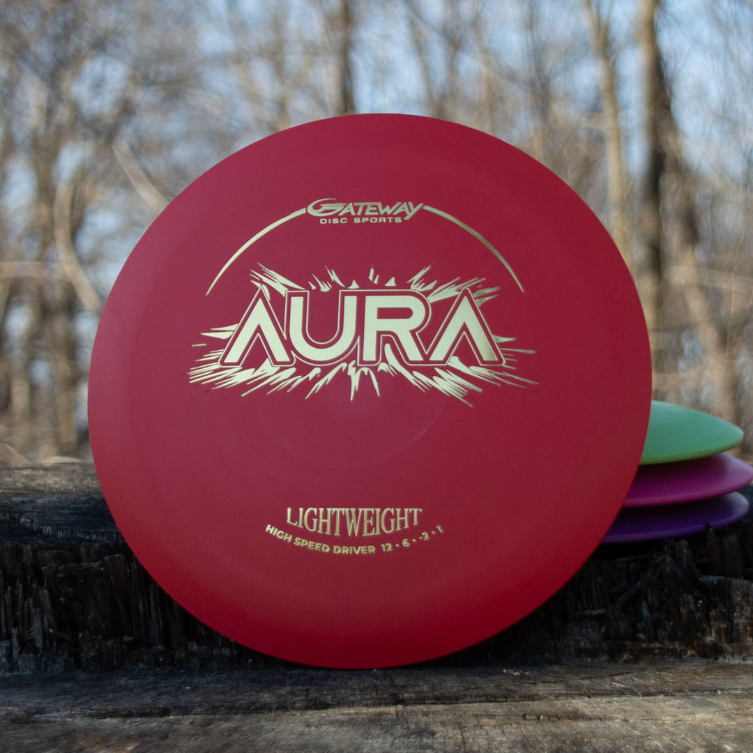 A red Aura - Suregrip Lightweight driver labeled "LIGHTWEIGHT" sits on a tree stump among other colorful lightweight discs, offering easy turnover and full flights for your next disc golf round.