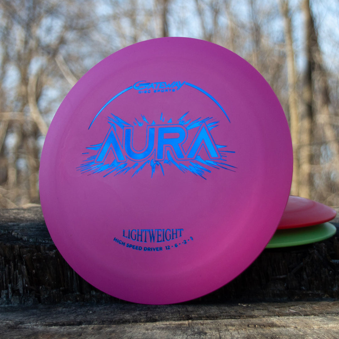 A pink Aura - Suregrip Lightweight disc golf driver stands upright on a wooden surface outdoors with trees behind it—ideal for easy turnover shots and full flights.