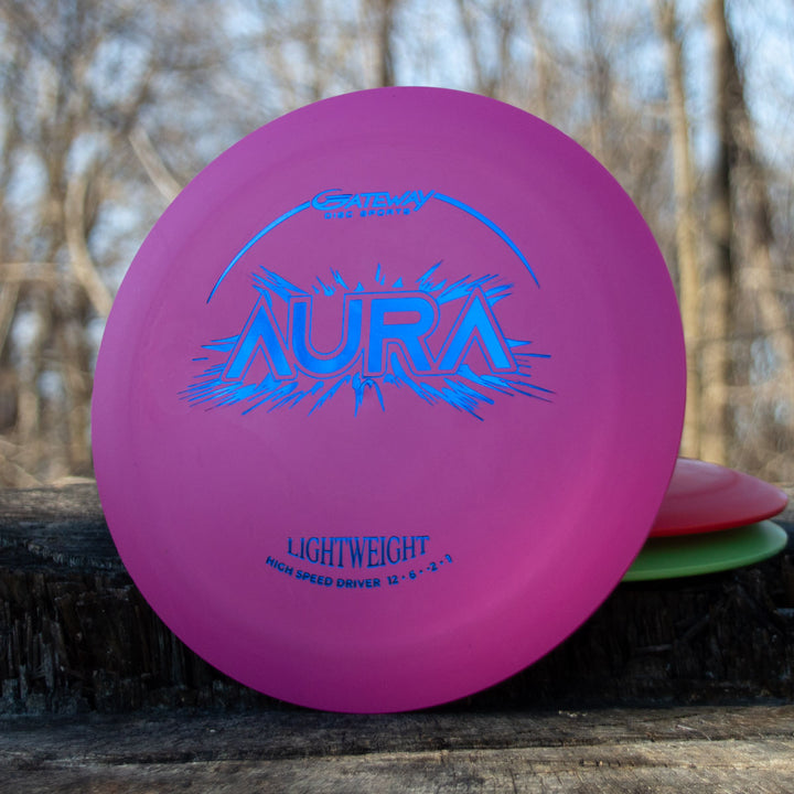 A pink Aura - Suregrip Lightweight disc golf driver stands upright on a wooden surface outdoors with trees behind it—ideal for easy turnover shots and full flights.