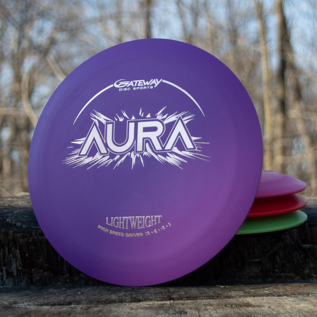 A purple Aura - Suregrip Lightweight disc golf driver labeled "LIGHTWEIGHT" rests against a stack of lightweight discs on a wooden surface outdoors.