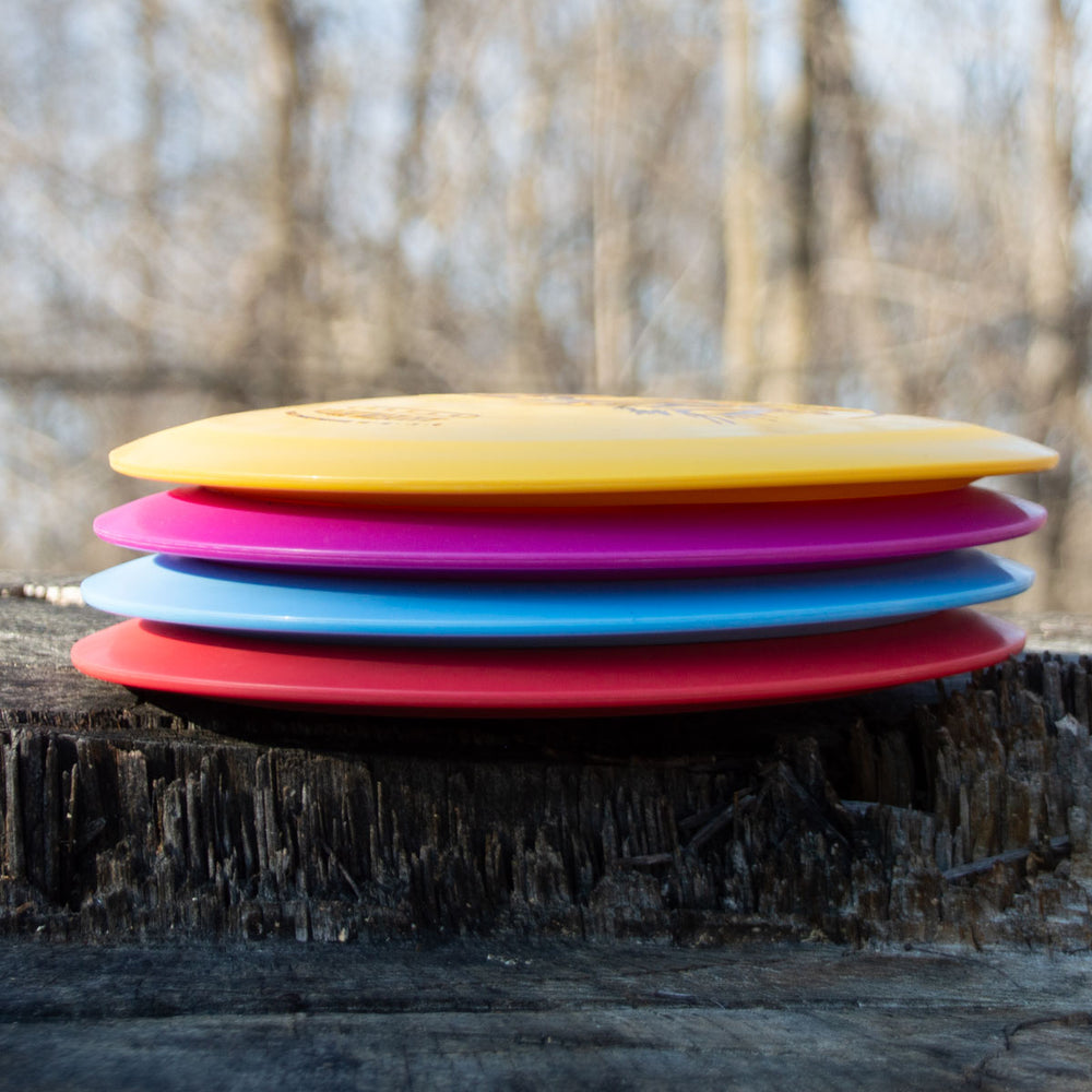 Four brightly colored Aura - Suregrip Ultra Lightweight discs are stacked on a tree stump outdoors, blurred leafless trees in the background—perfect for long flights on the course.