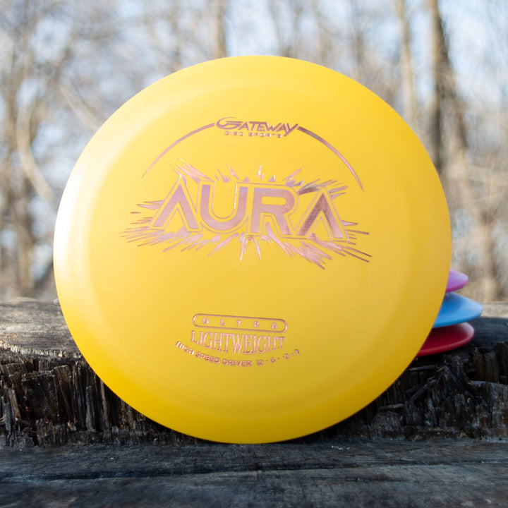An Aura - Suregrip Ultra Lightweight disc golf driver in yellow, designed for easy turnover and long flights, sits on a wooden surface outdoors with blurred trees in the background.