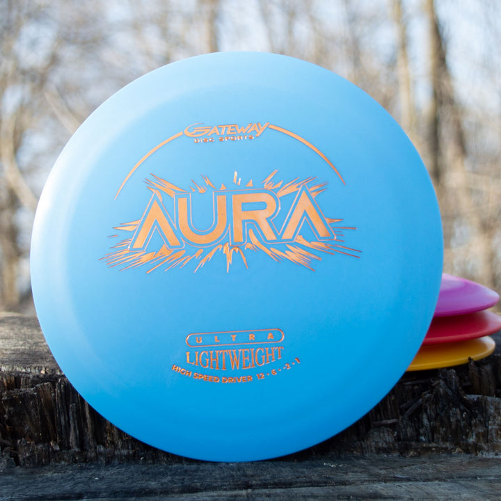 An Aura - Suregrip Ultra Lightweight disc golf driver sits on a wooden surface outdoors, its blue color standing out against blurred trees and other discs in the background, highlighting its easy turnover and long flight capabilities.