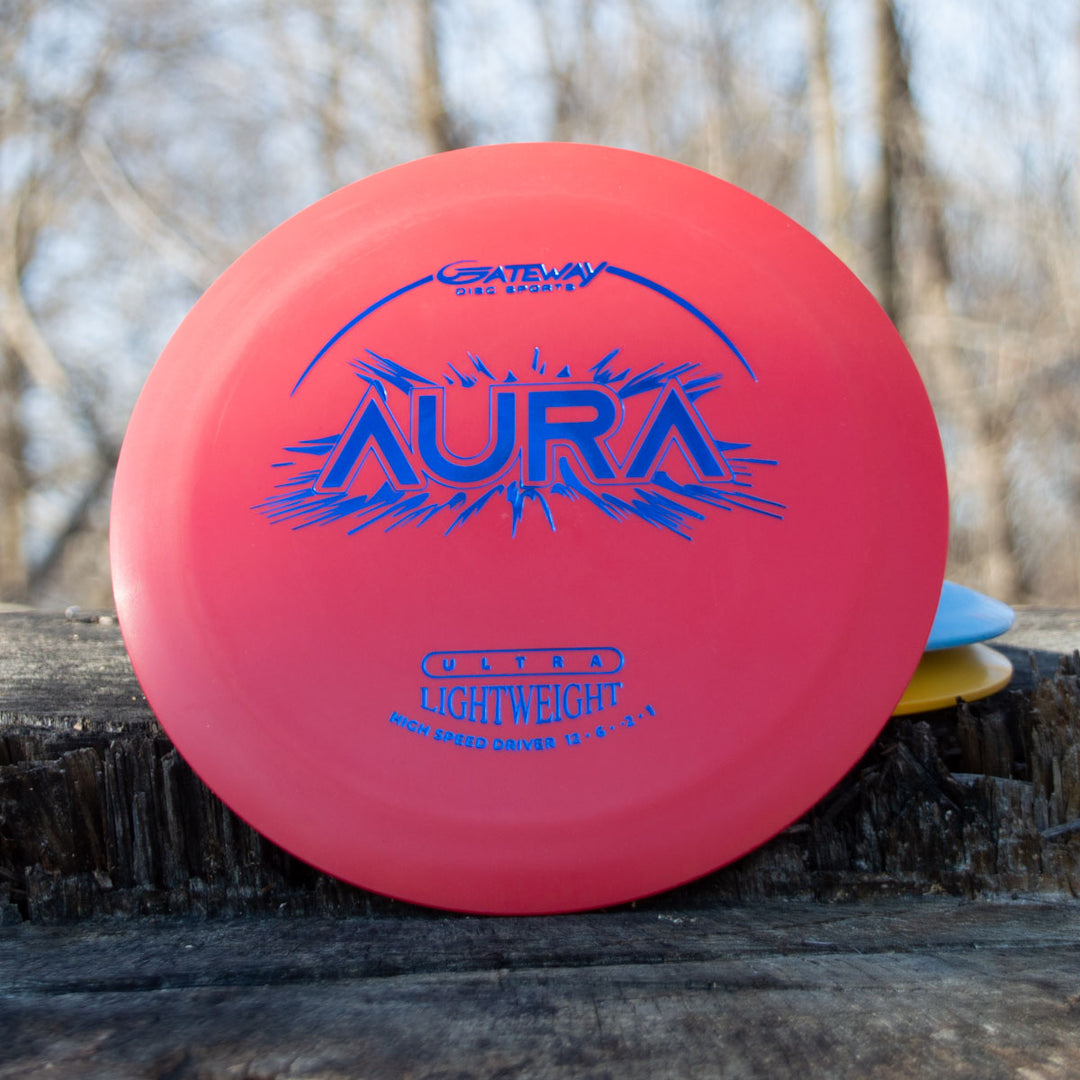 A red Aura - Suregrip Ultra Lightweight disc golf disc, designed for easy turnover and long flights, lies on a wooden surface outdoors with blurred trees in the background.