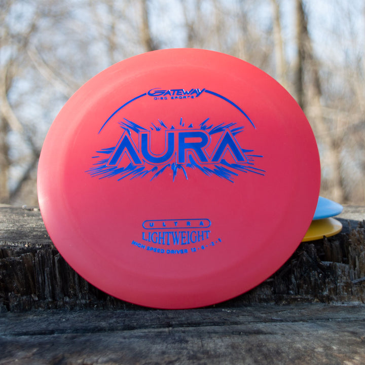 A red Aura - Suregrip Ultra Lightweight disc golf disc, designed for easy turnover and long flights, lies on a wooden surface outdoors with blurred trees in the background.