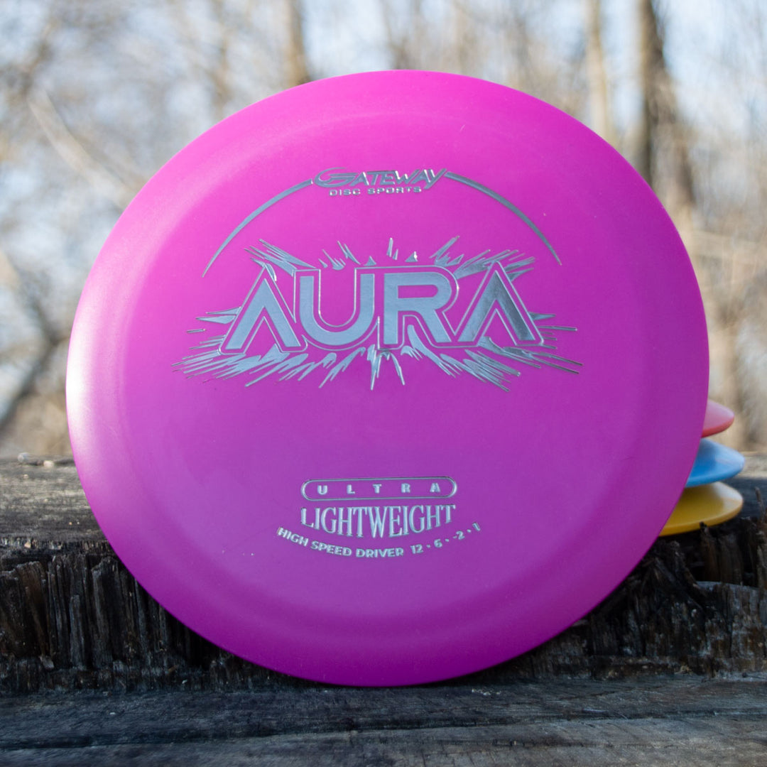 The Aura - Suregrip Ultra Lightweight, a pink disc golf disc with silver text and graphics, sits upright on a wooden surface outdoors with blurred trees in the background.