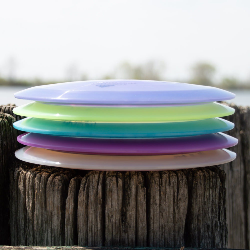 A stack of five colorful Aura - Diamond disc golf discs, featuring a less-stable model renowned for easy turnover, rests horizontally on a weathered wooden post with a blurred outdoor background.