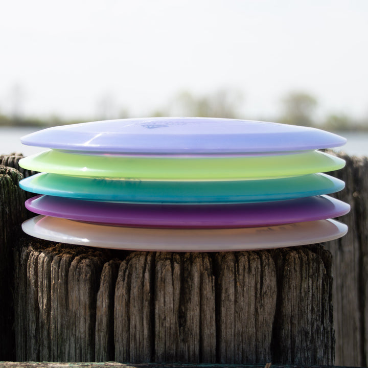A stack of five colorful Aura - Diamond disc golf discs, featuring a less-stable model renowned for easy turnover, rests horizontally on a weathered wooden post with a blurred outdoor background.