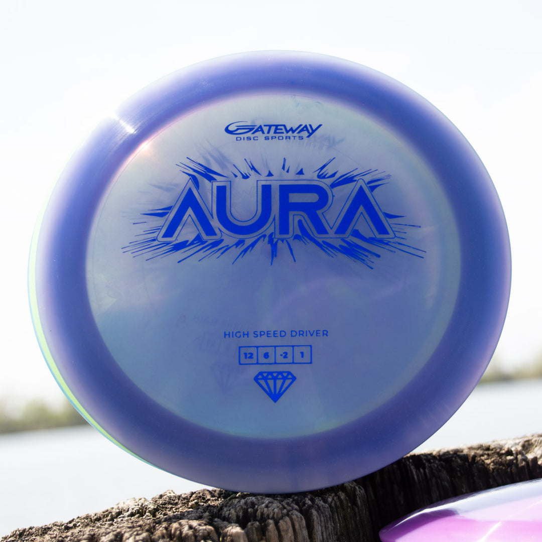 A blue Aura - Diamond high-speed driver disc from Gateway Disc Sports, known for its easy turnover, rests on a wooden surface outdoors near water.