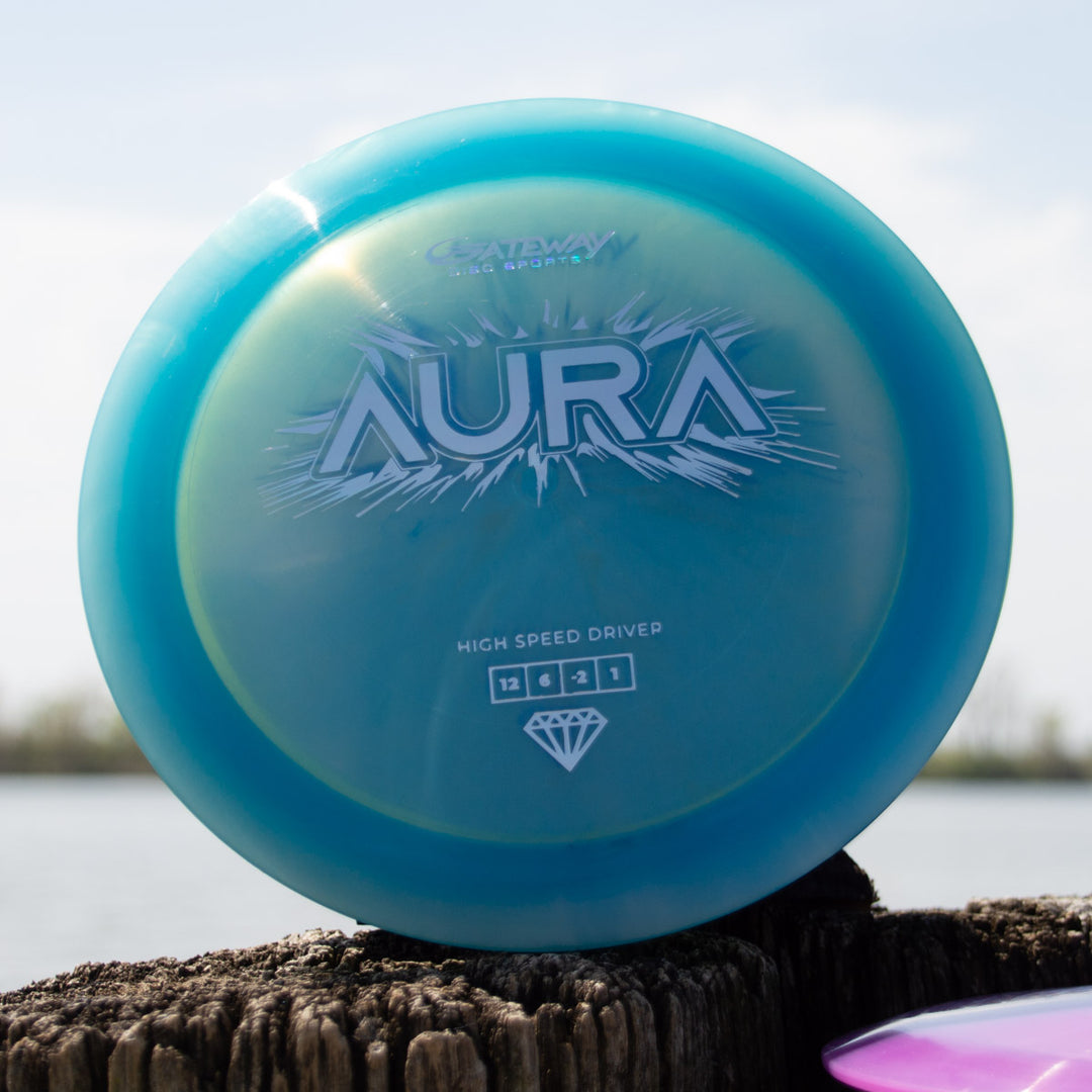 The Aura - Diamond is a blue high-speed driver disc golf disc with white text, recognized for its easy turnover ability, shown upright on a wooden post by the water.