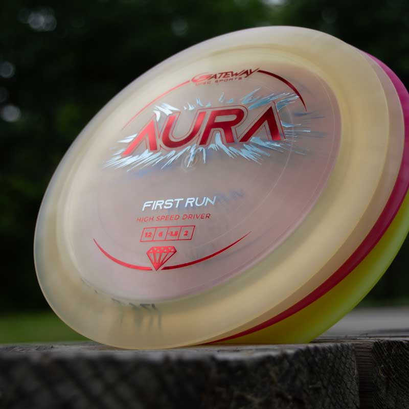 Close-up of an Aura - Diamond disc golf driver, known for easy turnovers, featuring "First Run" and "High Speed Driver" text, resting on a wooden surface outdoors.