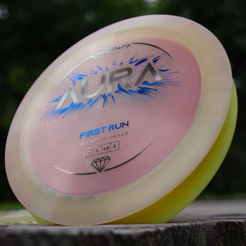 A close-up of the Aura - Diamond disc golf driver with a pink center and yellow rim, known for its long full flights, resting on a wooden surface outdoors.