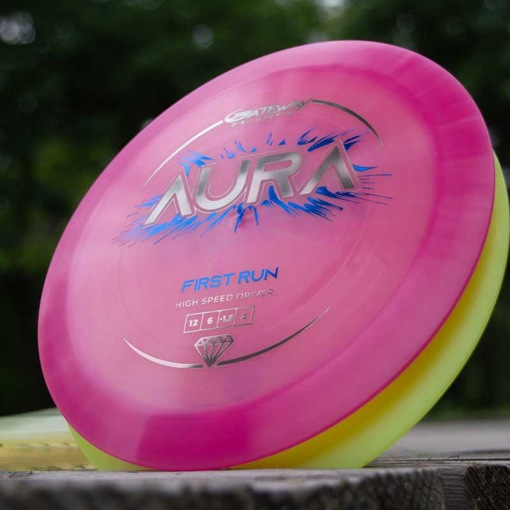 A close-up of the "Aura - Diamond" disc golf driver in pink and yellow, labeled "First Run" and known for its easy turnover, rests on a wooden surface outdoors.