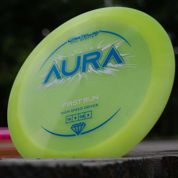 A yellow-green disc golf driver called "Aura - Diamond" from Gateway Disc Sports, known for easy turnovers and long flights, rests on a wooden surface with a blurred background.