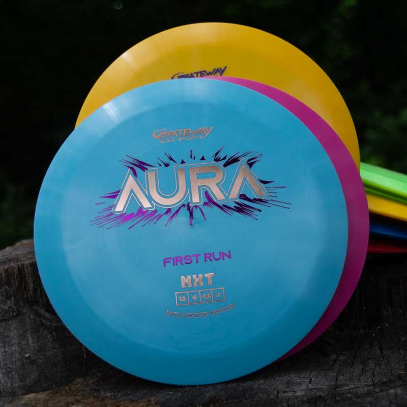 A blue Aura - NXT disc golf driver, prized for its turnover ability and long flights, sits on a tree stump in front of a yellow disc and several others.