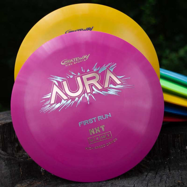 A close-up of the pink Aura - NXT disc golf disc in front of a yellow disc, both upright with more discs stacked behind, highlighting the turnover ability valued by players seeking long flights.
