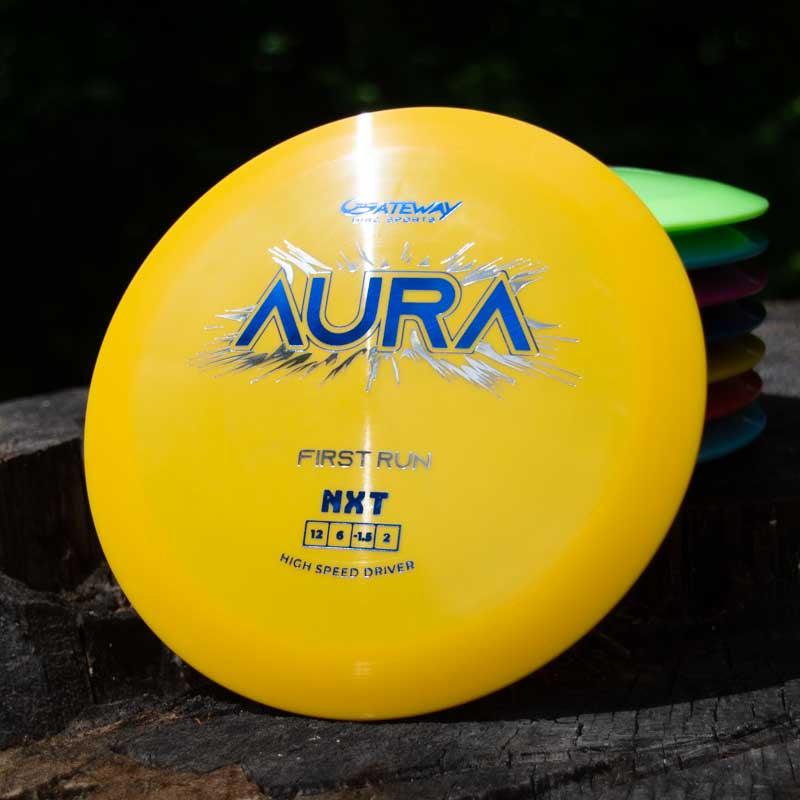 A yellow Gateway Disc Sports Aura - NXT disc golf driver labeled "FIRST RUN" and "HXT" rests on a tree stump, promising long flights, with other discs partially visible in the background.