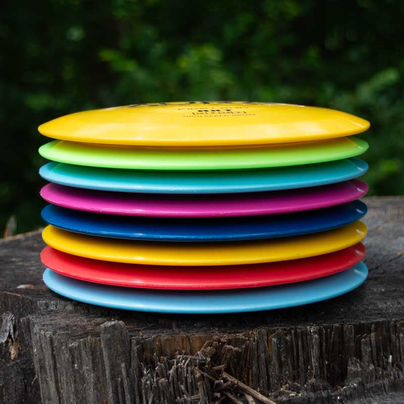 A stack of Aura - NXT disc golf discs, recognized for their turnover ability and long flights, sits on a tree stump with a blurred green background.