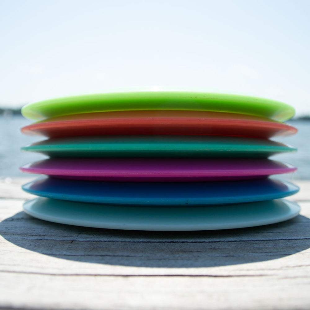 Six Aura - NXT Lite disc golf discs are stacked horizontally outdoors with water and sky in the background, highlighting their Nylon Extra Tough durability.