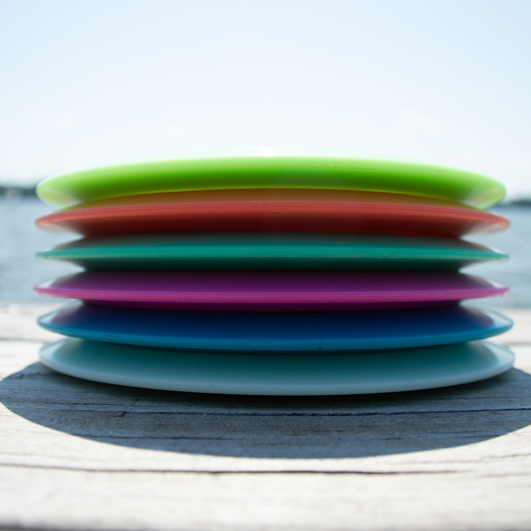 Six Aura - NXT Lite disc golf discs are stacked horizontally outdoors with water and sky in the background, highlighting their Nylon Extra Tough durability.