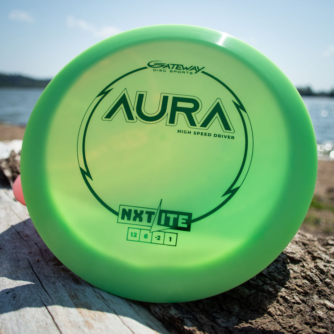 A green Aura - NXT Lite high-speed driver disc by Gateway Disc Sports sits on a log outdoors with a lake and sky in the background, crafted from Nylon Extra Tough plastic for added durability.
