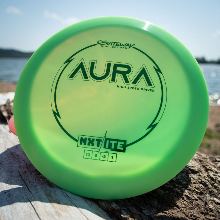 A green Aura - NXT Lite high-speed driver disc by Gateway Disc Sports sits on a log outdoors with a lake and sky in the background, crafted from Nylon Extra Tough plastic for added durability.