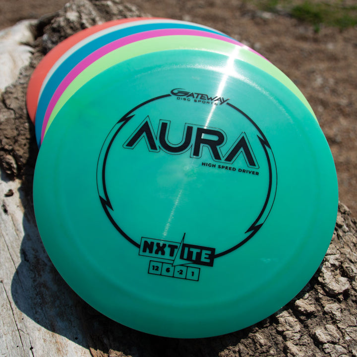 Four colorful Aura - NXT Lite high-speed disc golf discs are stacked on a tree trunk, with the top disc showing the brand and model.