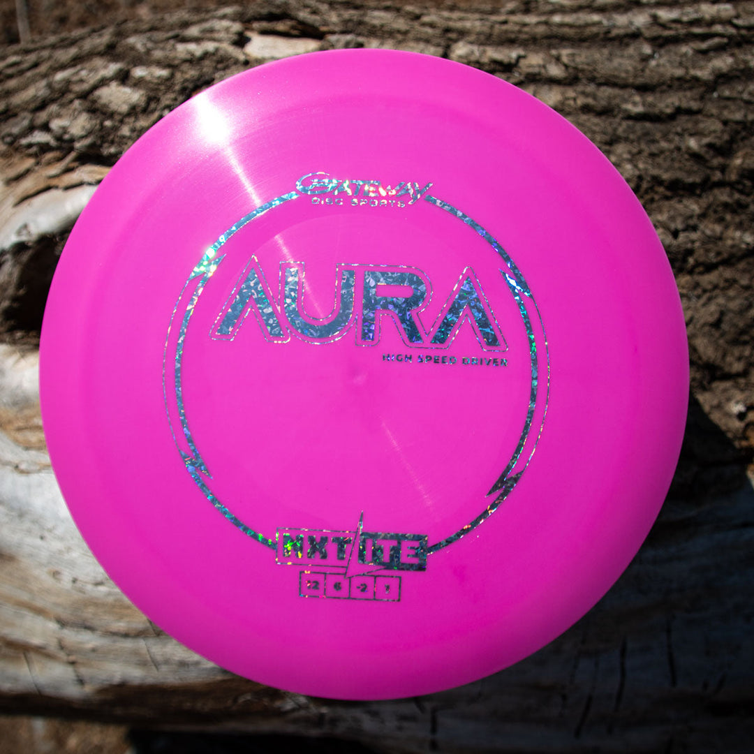 A pink Aura - NXT Lite disc golf disc made from Nylon Extra Tough material sits on a weathered, rough wooden surface outdoors.