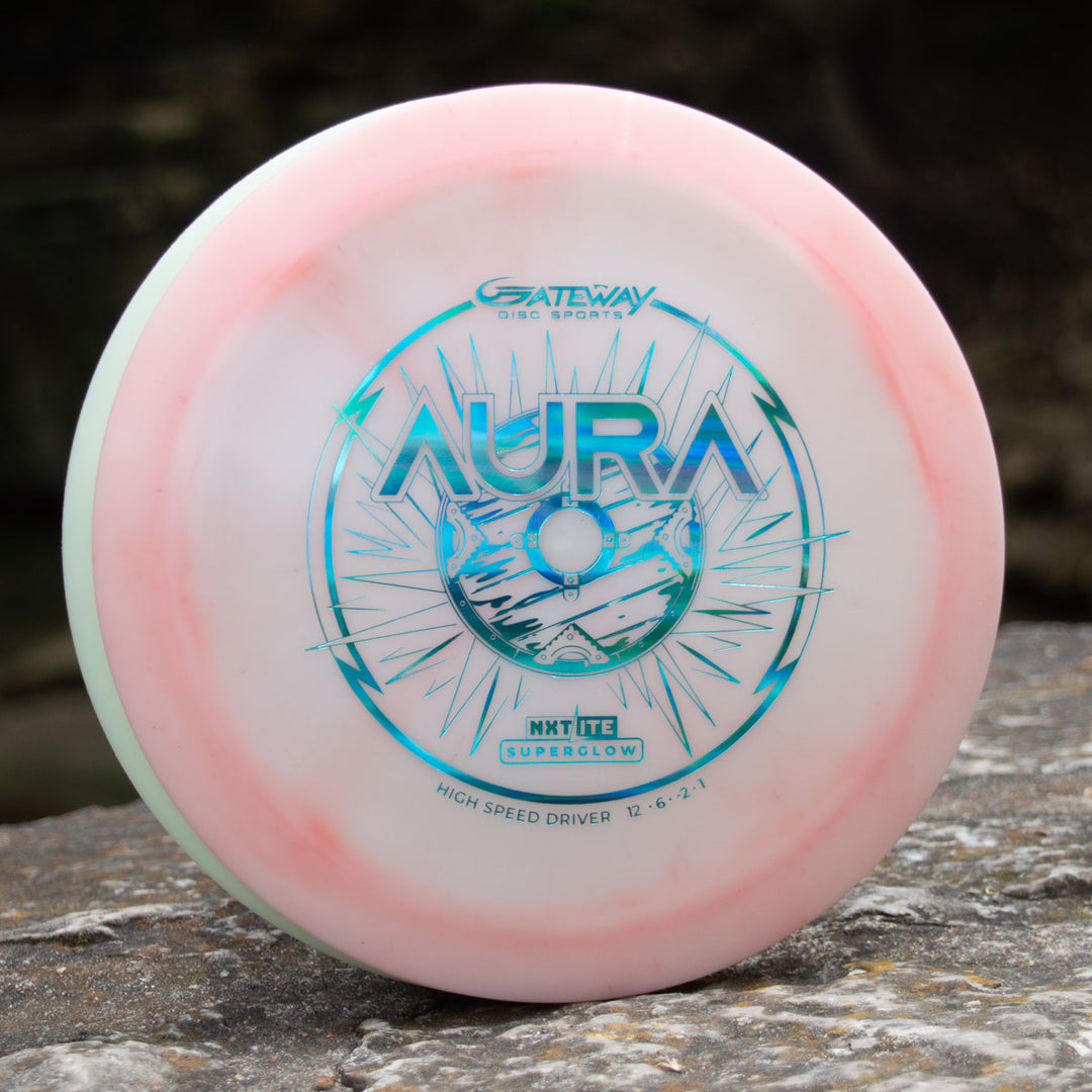 The Aura - NXT Lite Superglow, a white and light pink premium driver with blue artwork and "AURA" plus "GATEWAY DISC SPORTS" text, rests on a rock outdoors—ideal for smooth hyzer flips and ultimate glide.